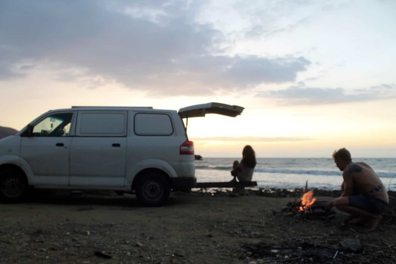 A couple make a fire on the beach next to a van