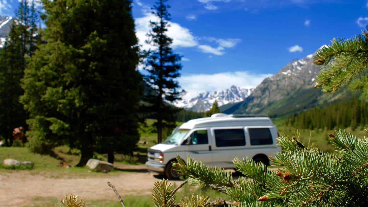 A van in the mountains in the middle ground. Snow-capped peaks behind, pine needles in focus. Van Life or Boat Life